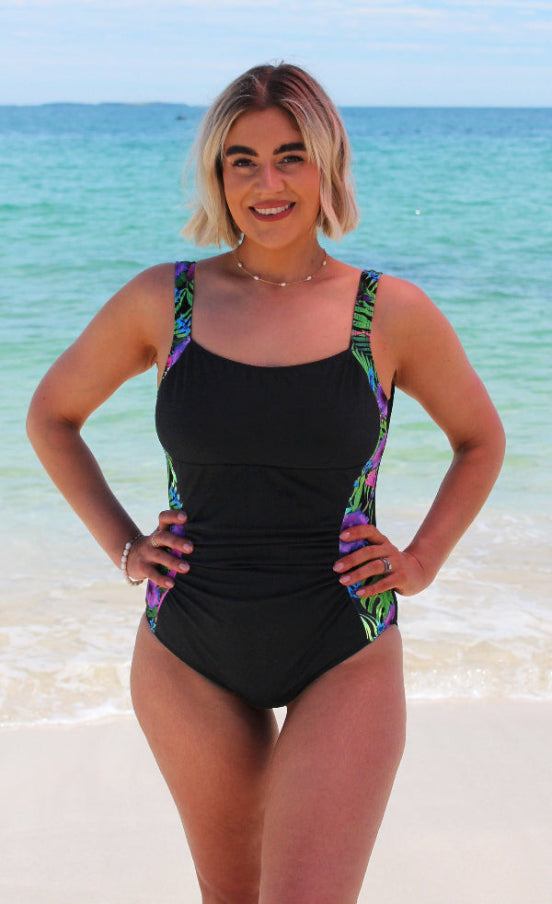 Woman in a black swimsuit with colorful straps standing on a beach.